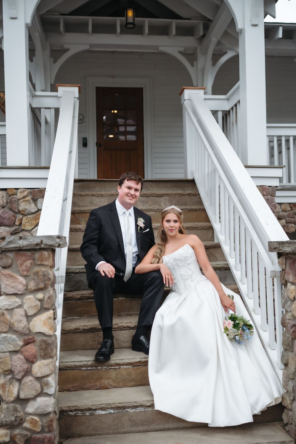Bride and groom sitting on stone steps of white Victorian porch in wedding attire.