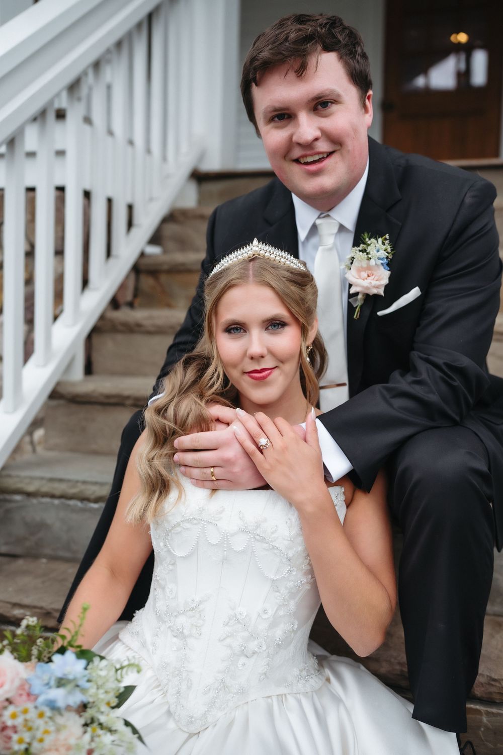 A smiling bride in a white gown and tiara sits on steps with a groom in a black suit behind her.