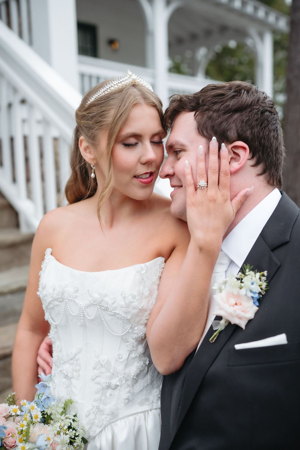 Bride in white floral gown with tiara tenderly touches groom's face in dark suit with boutonniere outside white venue.