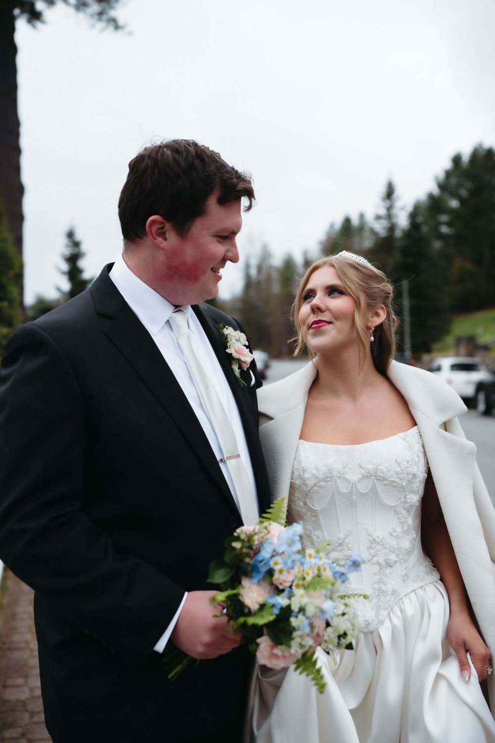 Bride and groom share a loving glance outdoors, bride in white coat holding blue bouquet, groom in black suit.