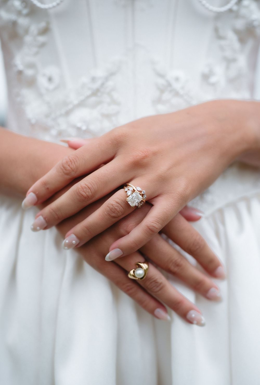 Bride wearing elegant diamond and pearl rings on manicured hands, dressed in white lace wedding gown.