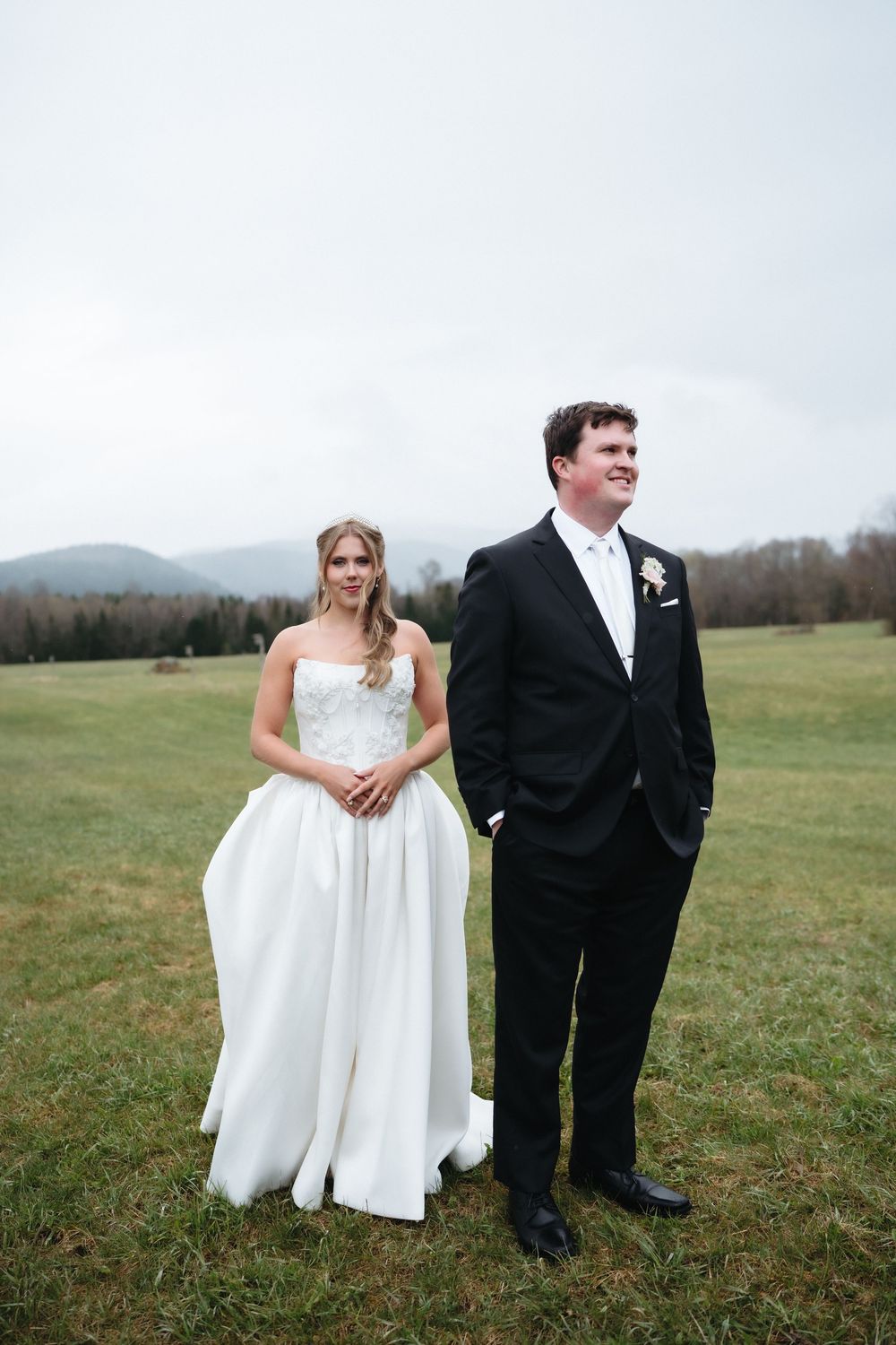 Bride in white strapless gown and groom in black tuxedo stand in green field with mountain backdrop.