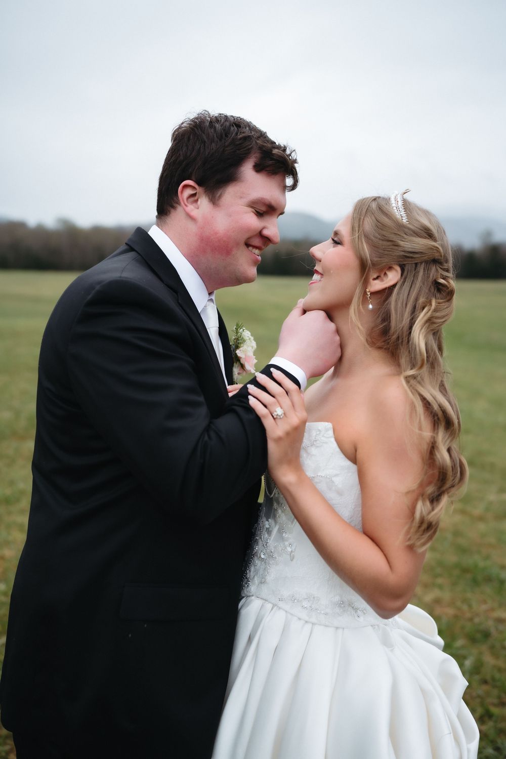 Bride and groom share a romantic moment outdoors, smiling at each other in wedding attire with green fields behind them.