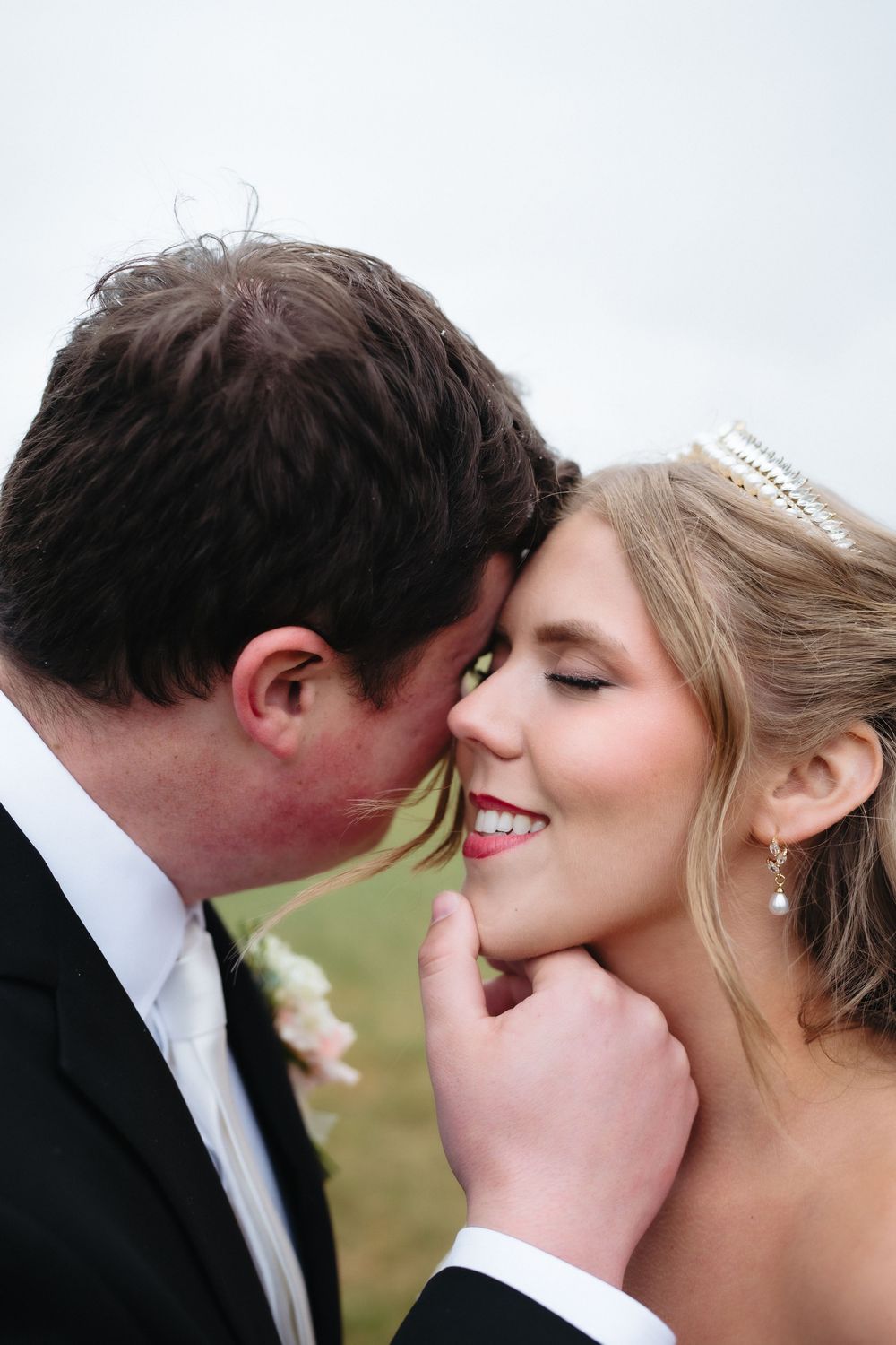 Groom tenderly kisses bride on cheek as she smiles, wearing pearl earrings and tiara on wedding day.