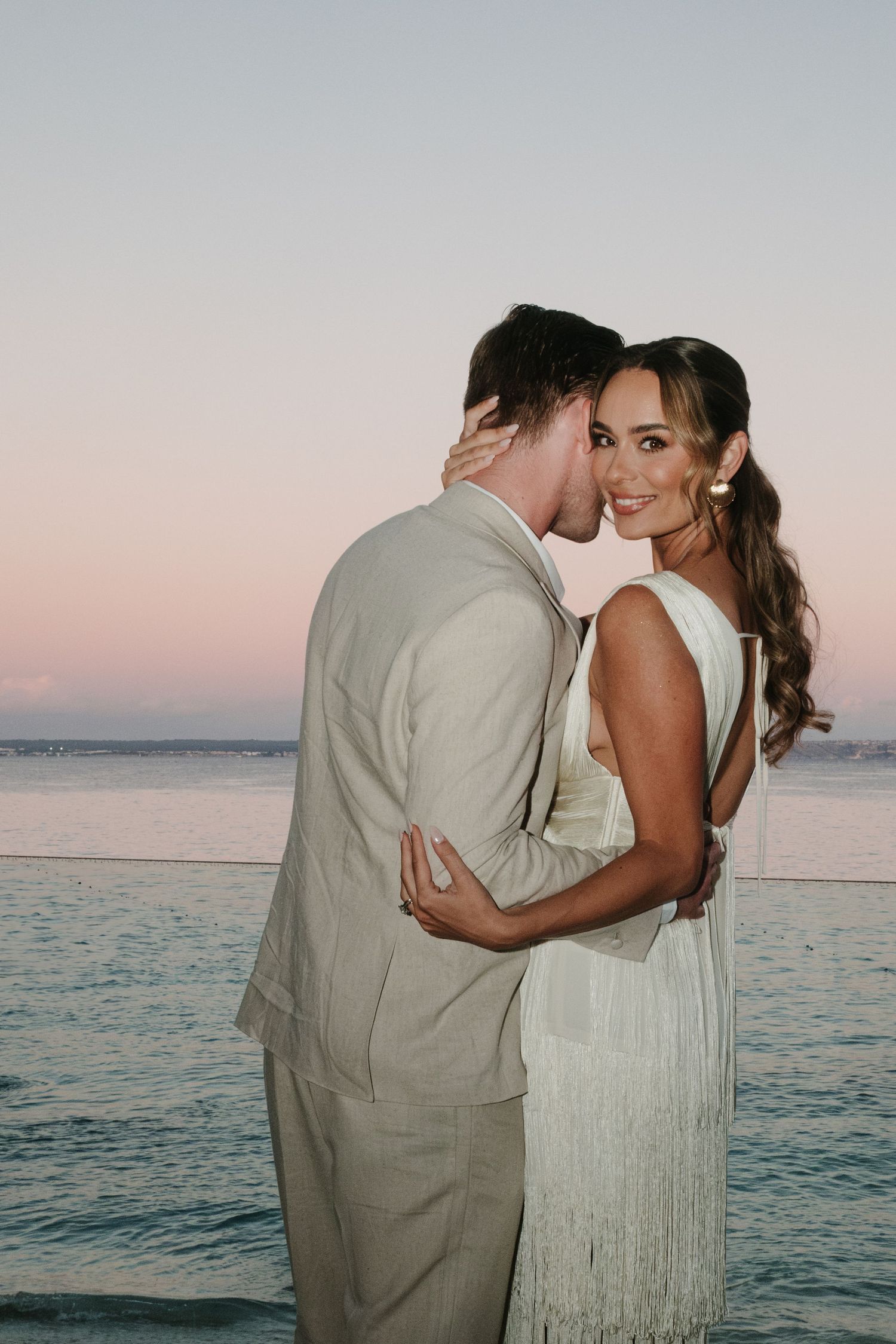 Couple embracing at sunset by the ocean, woman in white fringe dress and man in beige suit, romantic beach wedding photo.