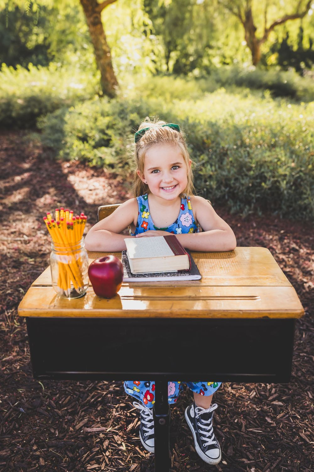 Young smiling girl sitting at a wooden school desk outdoors with pencils, apple, and books in a sunny garden setting.
