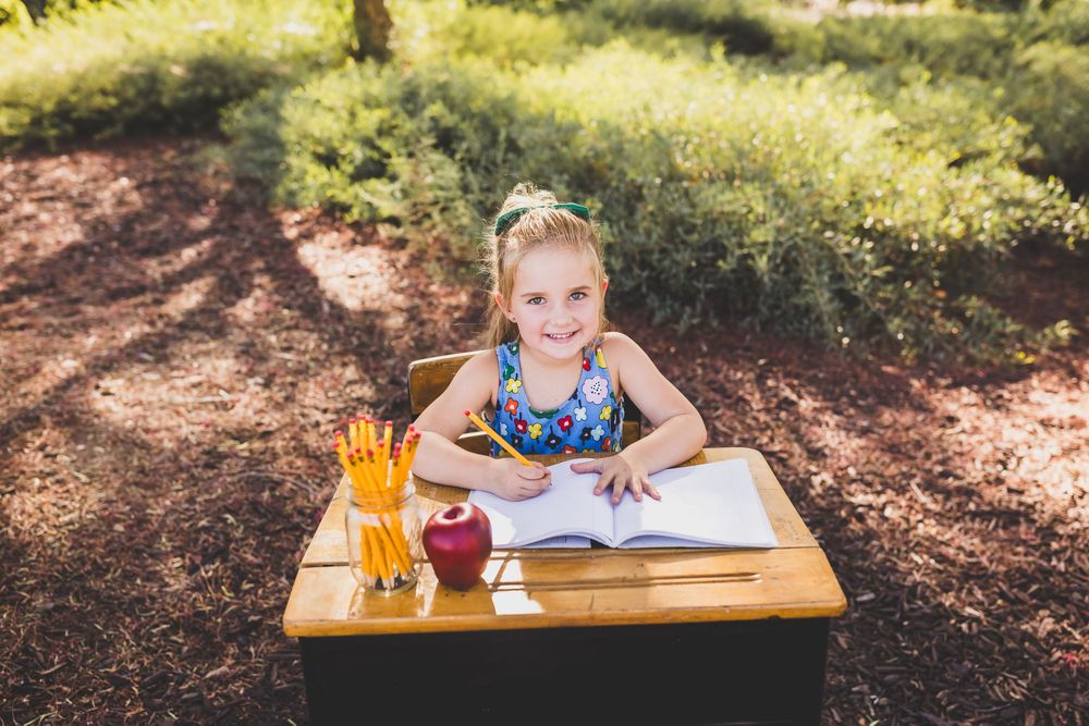 Young girl smiling at school desk outdoors with pencils, apple, and open notebook in sunny garden setting.
