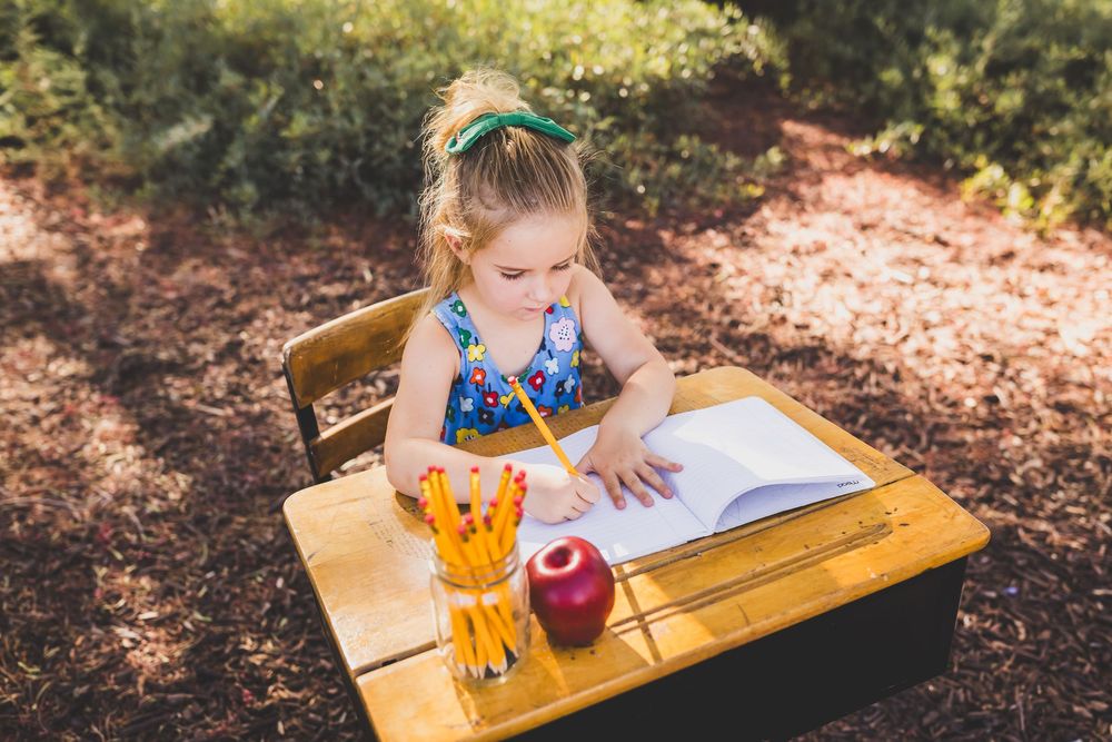 Young girl writing in notebook at wooden school desk outdoors with pencils and red apple, back to school concept.