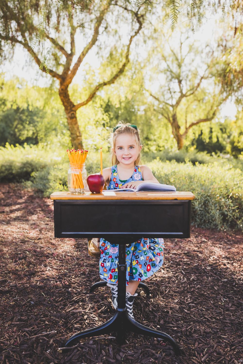 Young girl in floral dress sitting at vintage school desk outdoors, smiling with apple and pencils on desk.