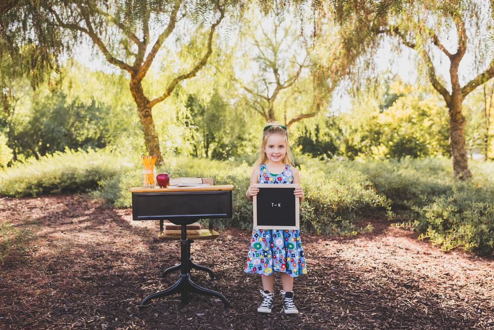 Young girl holding a chalkboard sign outdoors near a vintage desk, surrounded by trees in golden sunlight.