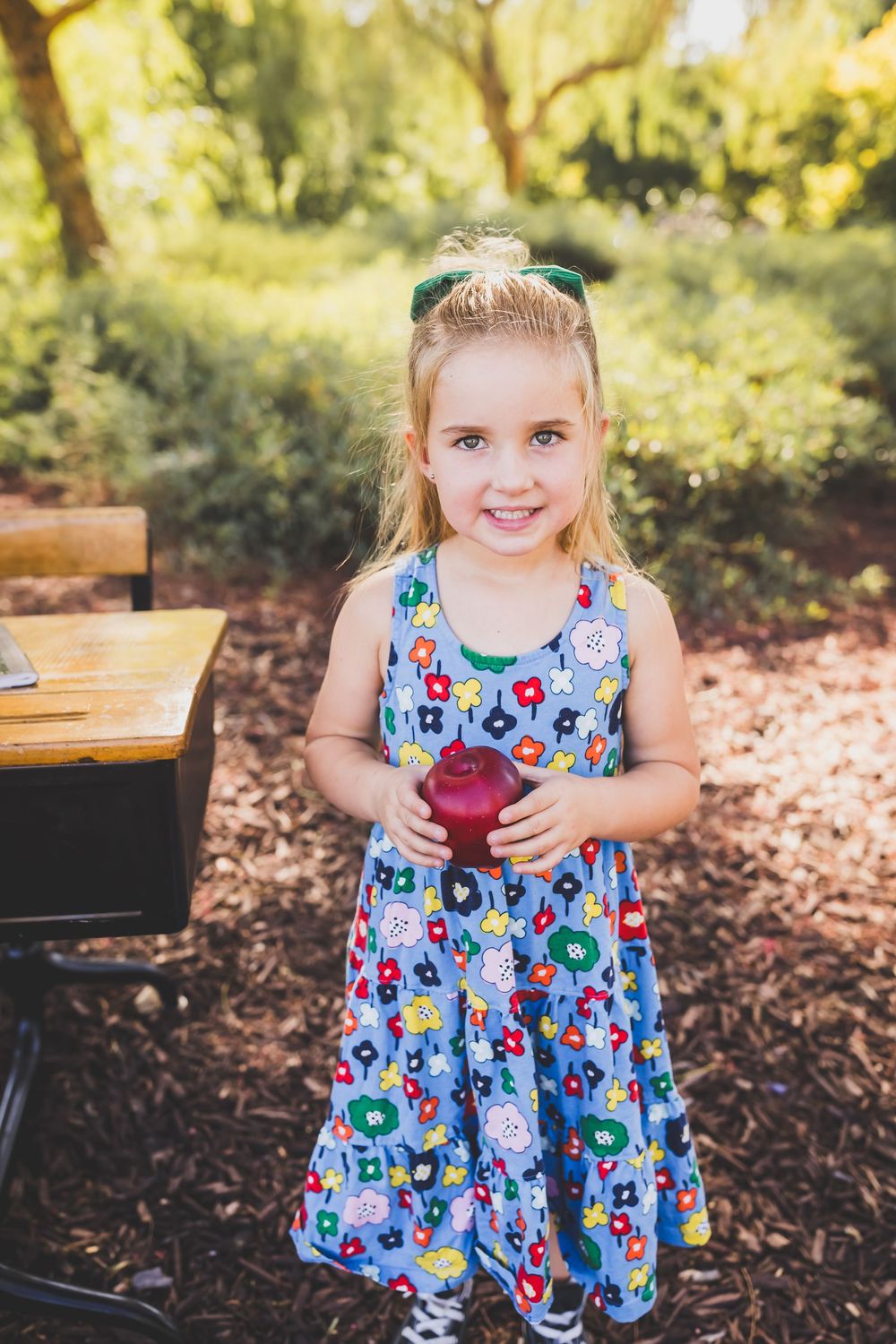 Young blonde girl in floral dress holding red apple outdoors near school desk, smiling in sunlit garden.