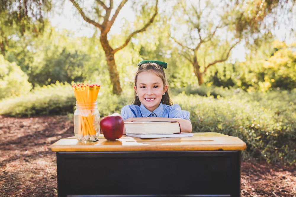 Smiling girl at outdoor school desk with pencils, apple, and books, surrounded by green trees.