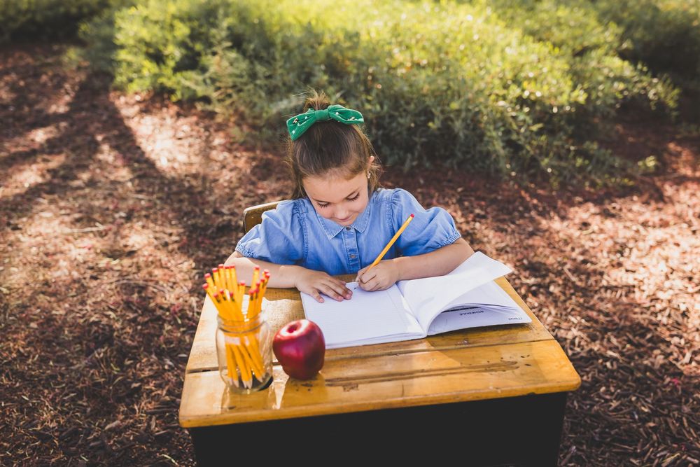 Young girl in blue dress writing in notebook at wooden desk outdoors, with pencils and red apple, in a school setting.