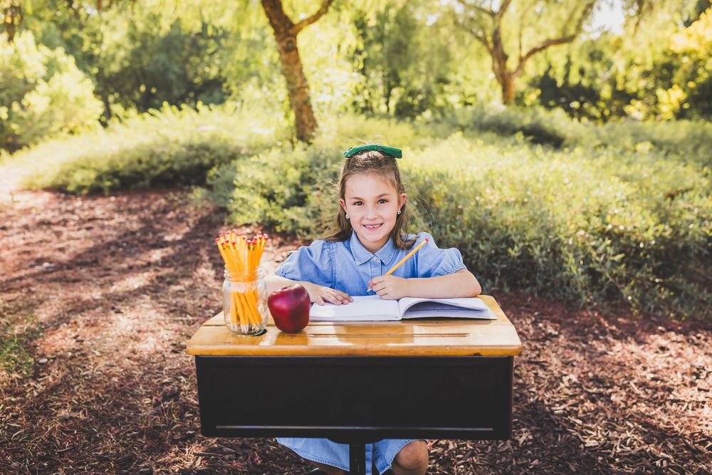 A smiling girl sits at an outdoor school desk with pencils, an apple, and an open book, surrounded by lush green trees.