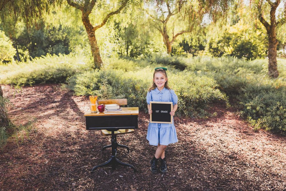 Girl in blue dress holds chalkboard sign beside vintage school desk outdoors among green trees on bark ground.