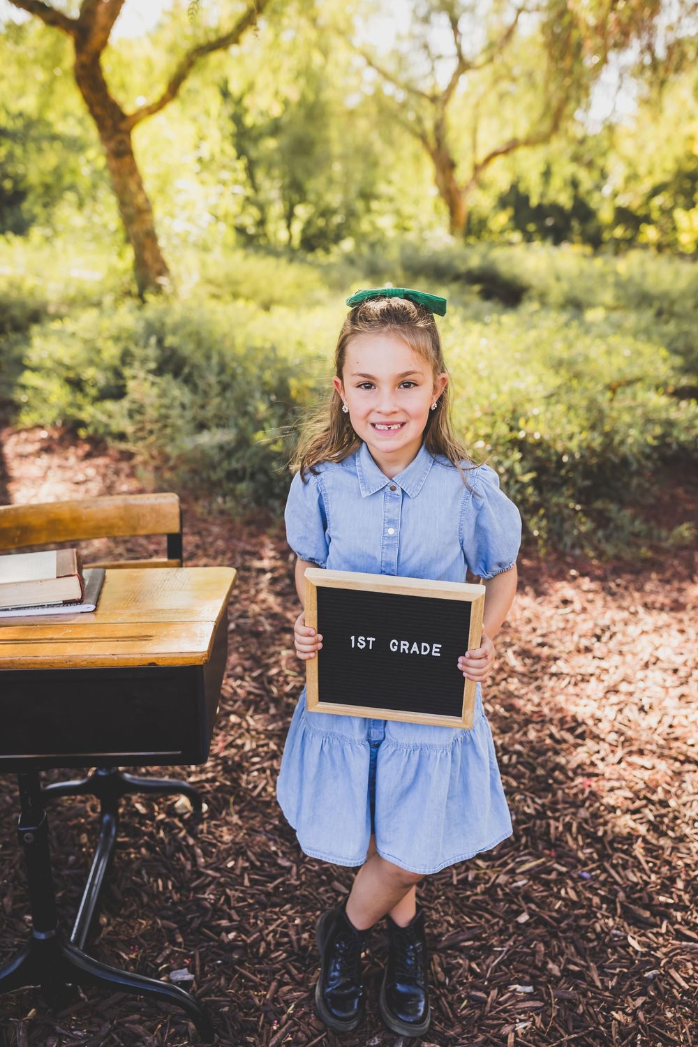 Smiling girl in blue dress holding a 1st Grade letterboard sign outdoors near a school desk among green trees.
