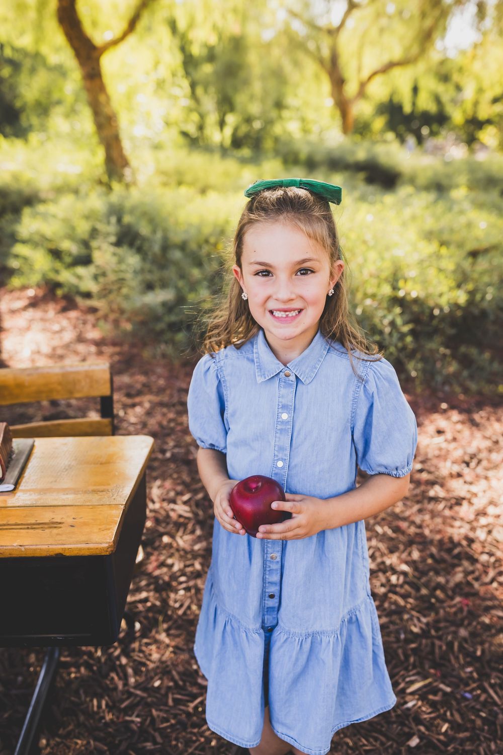 Smiling young girl in blue dress holding a red apple outdoors near a school desk among green trees.