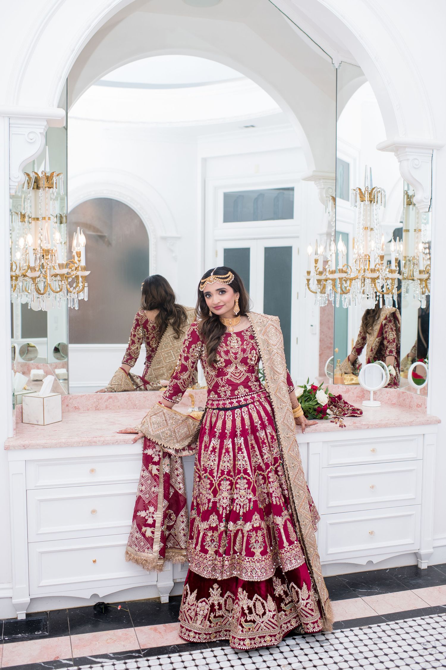 Bride in ornate red and gold lehenga poses in elegant white bathroom with chandelier and arched mirrors.