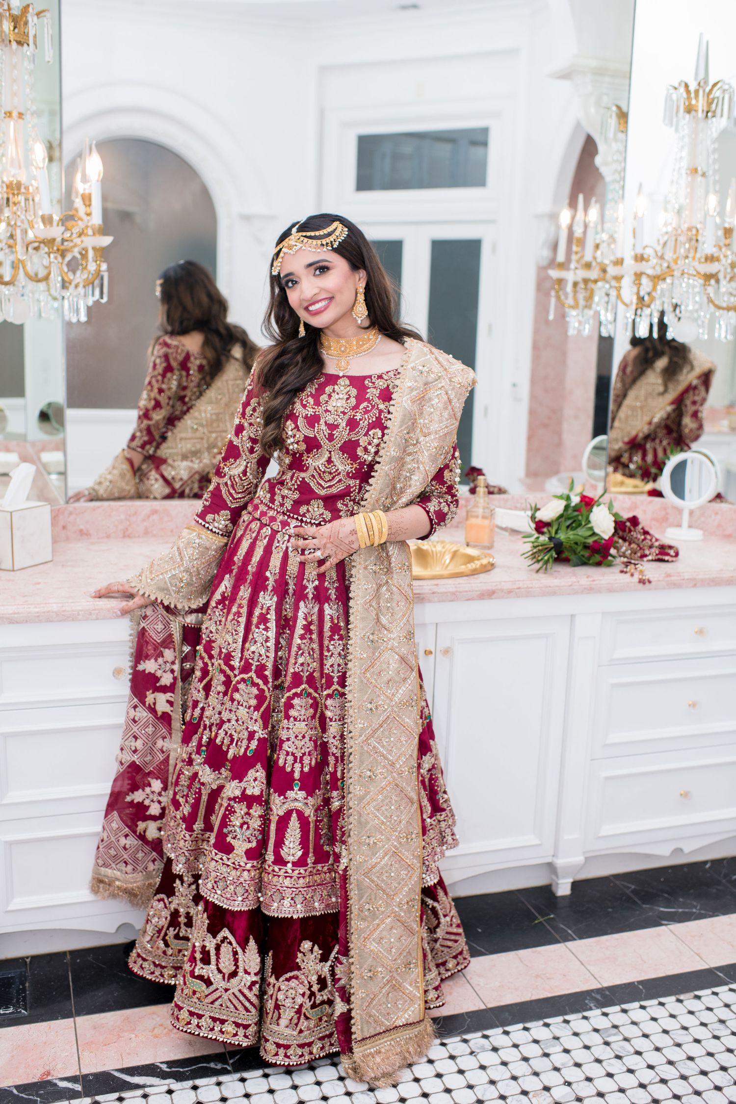 Bride in ornate crimson and gold embroidered lehenga with dupatta and maang tikka poses in elegant dressing room.
