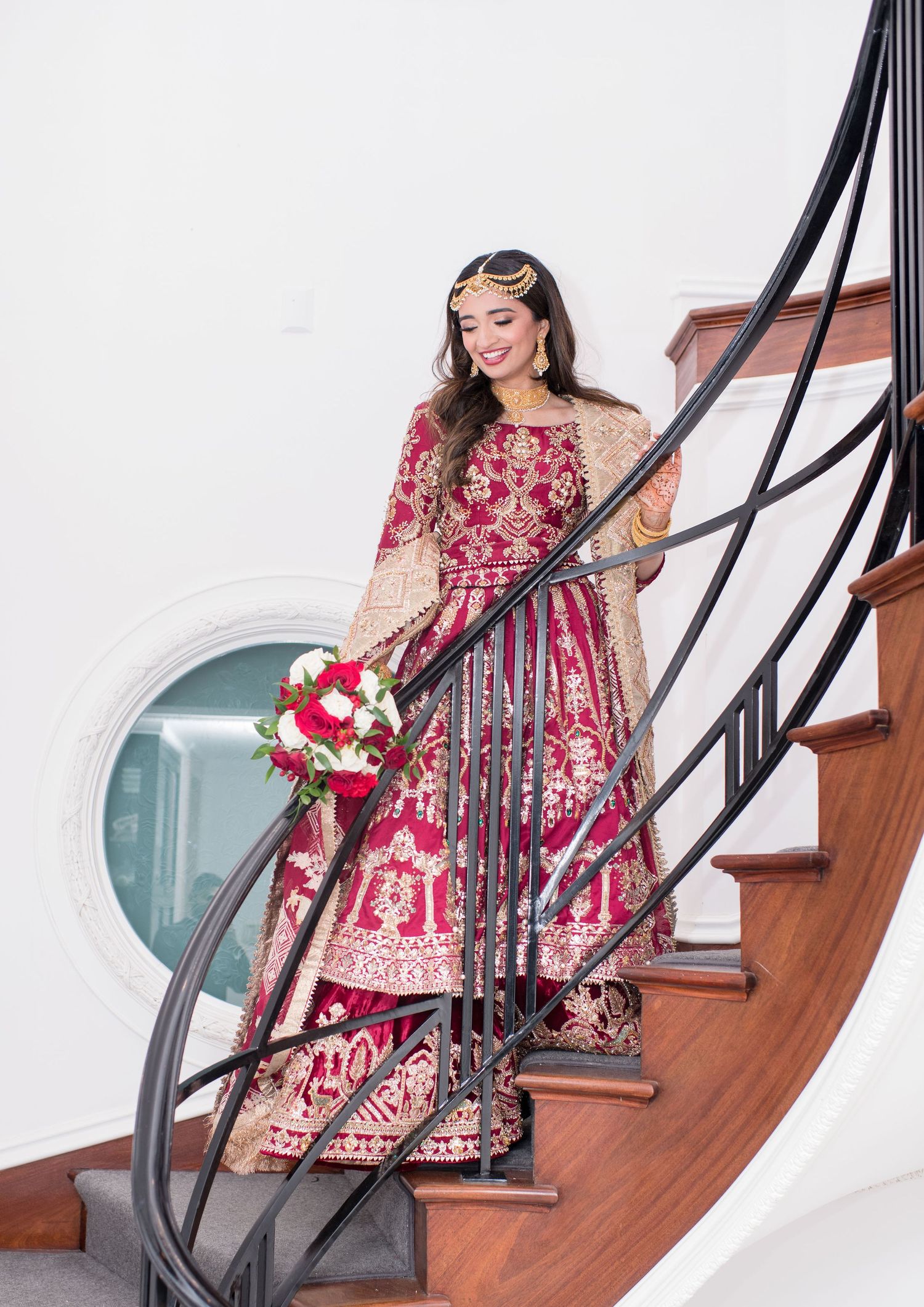 Bride in red and gold embroidered South Asian bridal gown holding bouquet on elegant curved staircase.