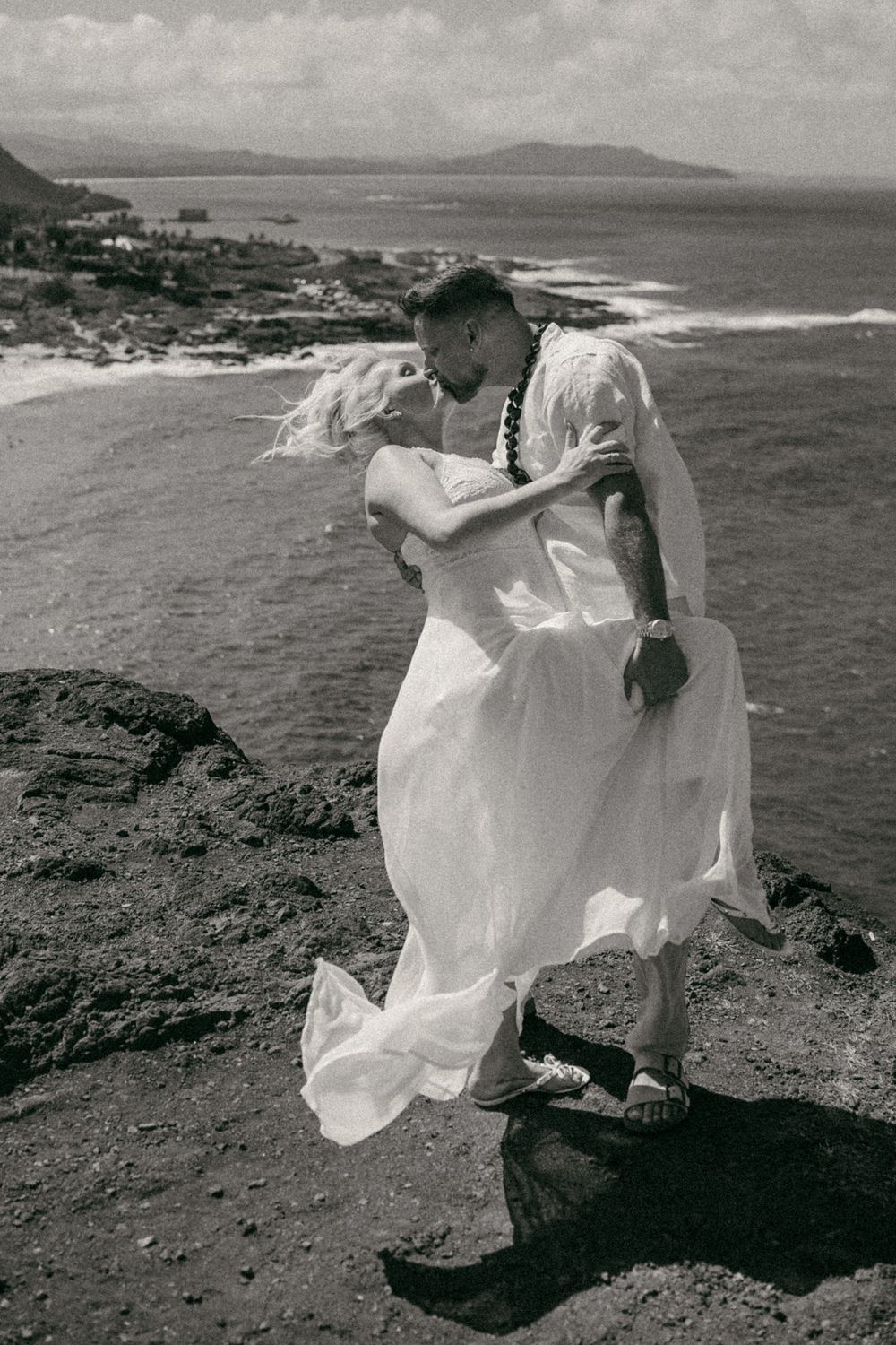 Black and white photo of a couple embracing on rocky cliffs overlooking the ocean, bride in flowing white dress.