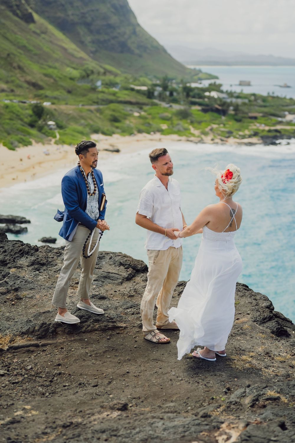 Couple exchanging vows during an outdoor beach wedding ceremony in Hawaii with an officiant on rocky cliffs.
