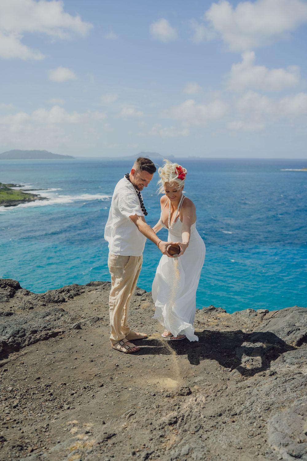 Couple in wedding attire performing sand ceremony on rocky cliff overlooking turquoise Hawaiian ocean waters.