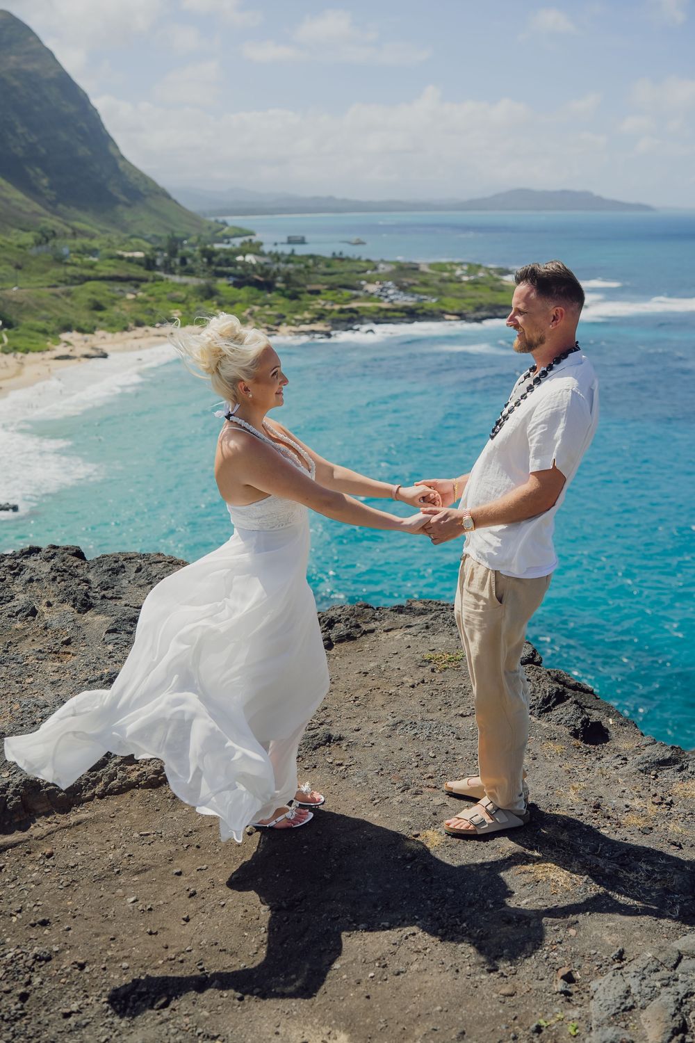 Couple holding hands in wedding attire on rocky cliff overlooking turquoise ocean in Hawaii.