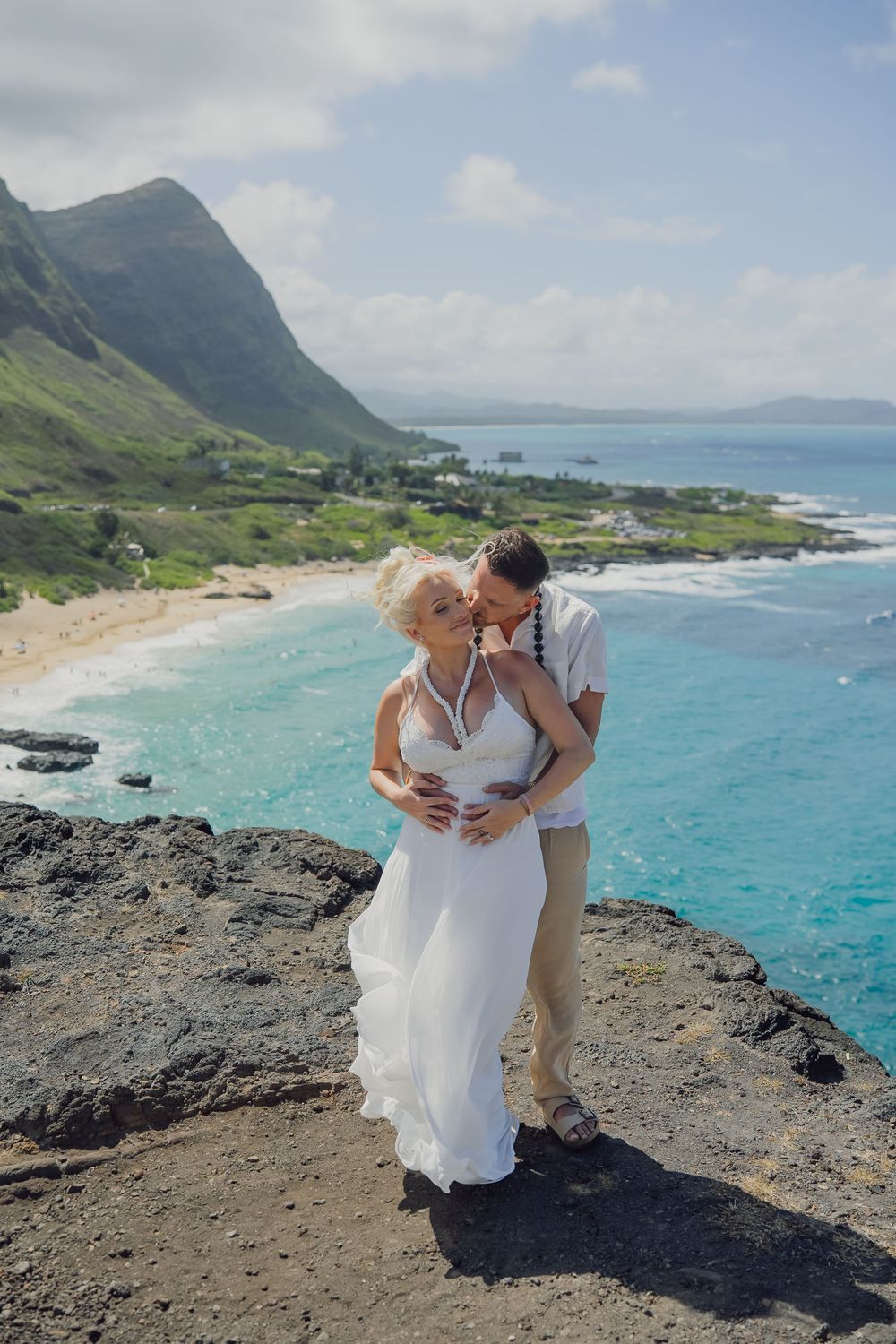 Couple in wedding attire embracing on rocky cliff overlooking turquoise ocean and green mountains in Hawaii.