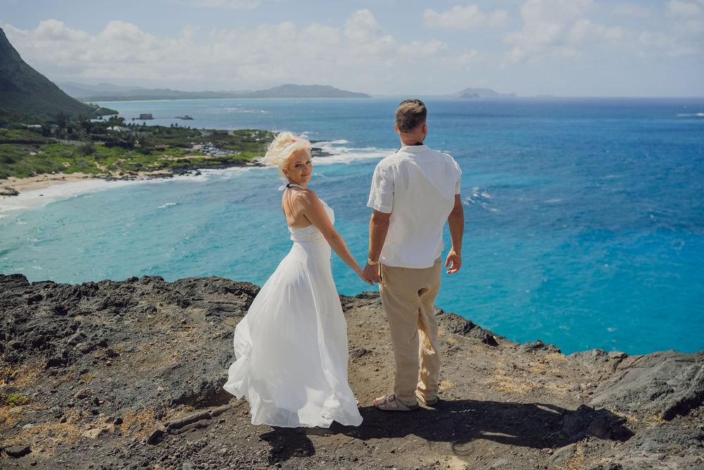 Bride and groom holding hands on rocky cliff overlooking stunning turquoise ocean and coastline in Hawaii.