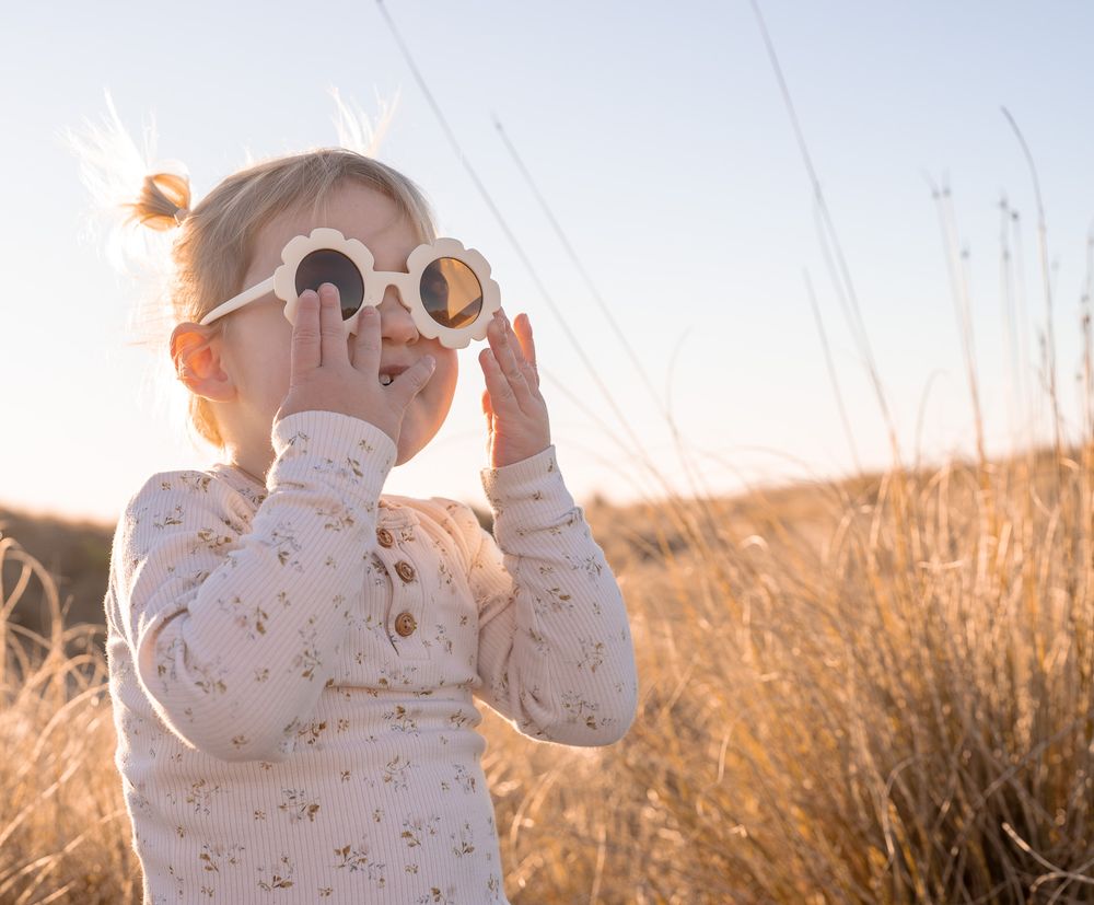 Toddler girl wearing white floral sunglasses in a golden wheat field during sunset.