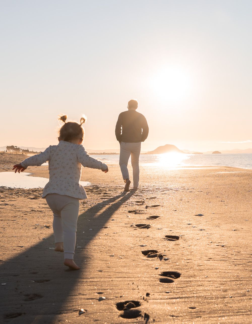 A child and adult run toward the sunset on a sandy beach, leaving footprints behind them.