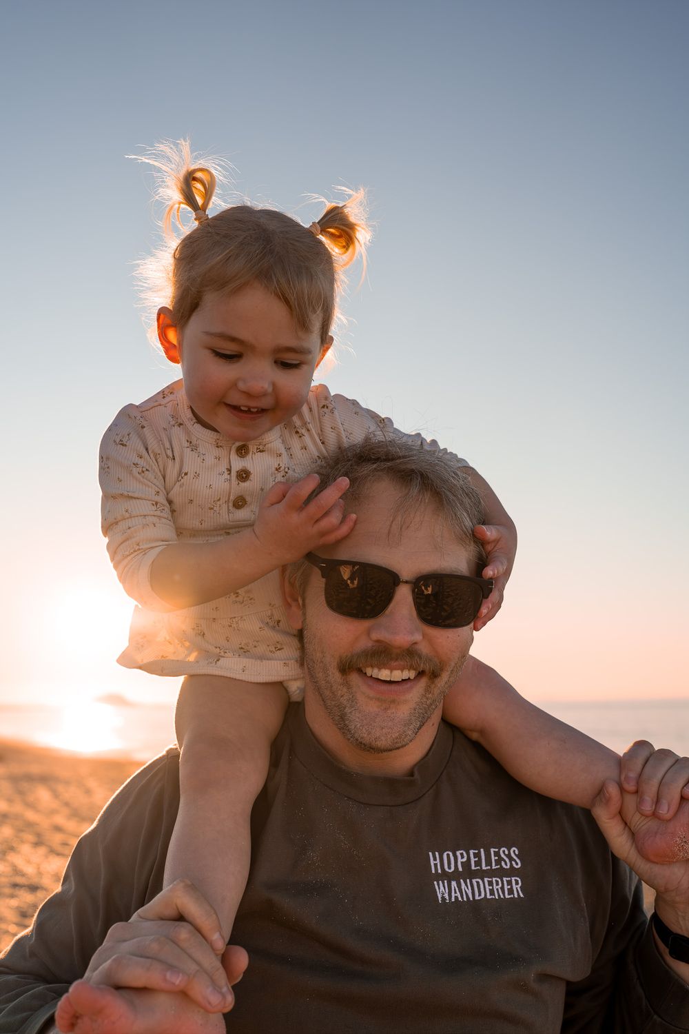 Happy toddler girl sitting on fathers shoulders at golden hour beach sunset.