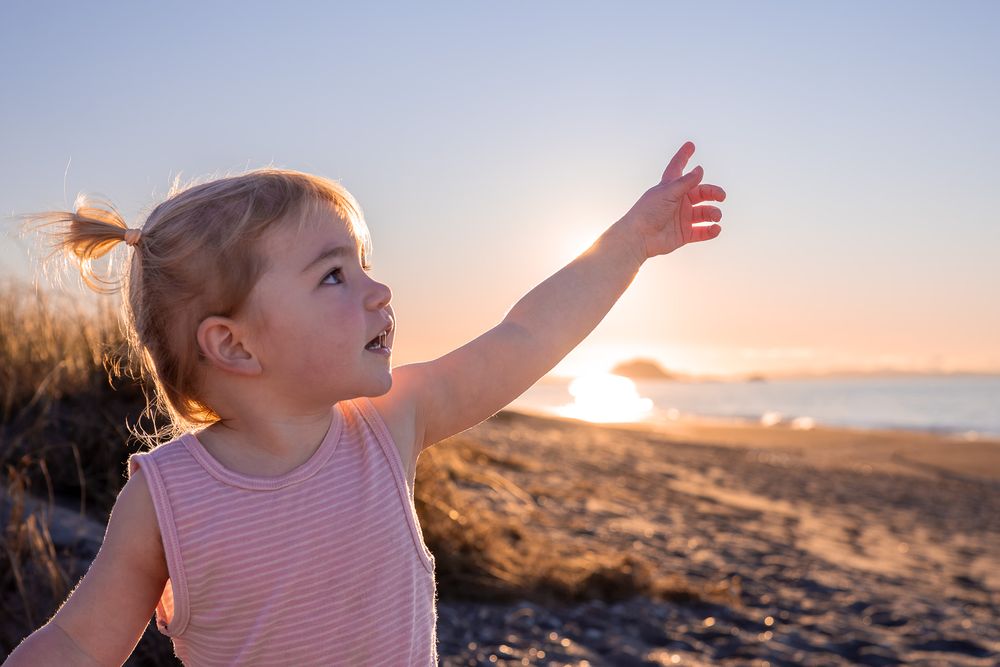 Curious toddler girl pointing at the horizon during a beautiful beach sunset.