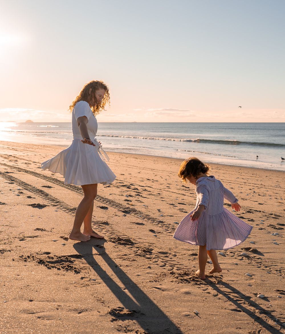 Mother and daughter in white dresses dancing on a sandy beach at golden sunset.