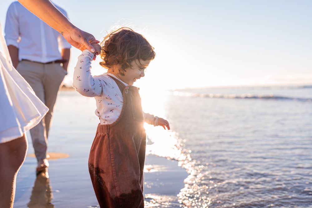 Young child walking on beach holding parent hand at sunset.