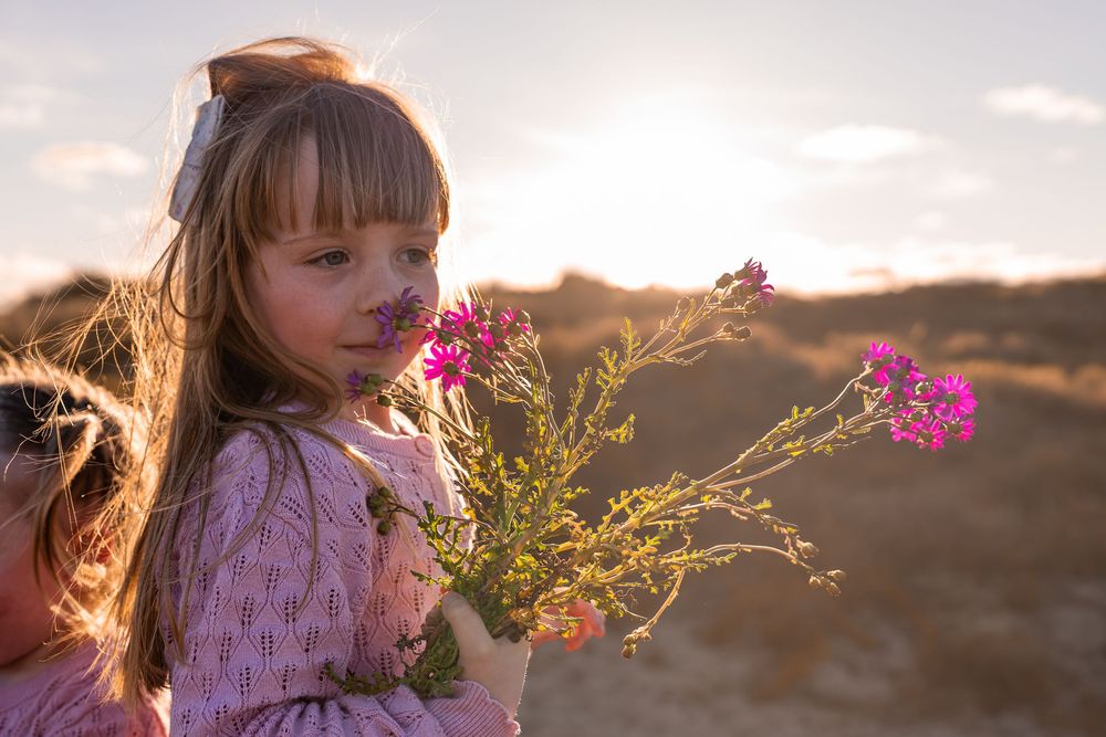 Young girl in pink dress holding pink wildflowers outdoors at golden sunset hour.
