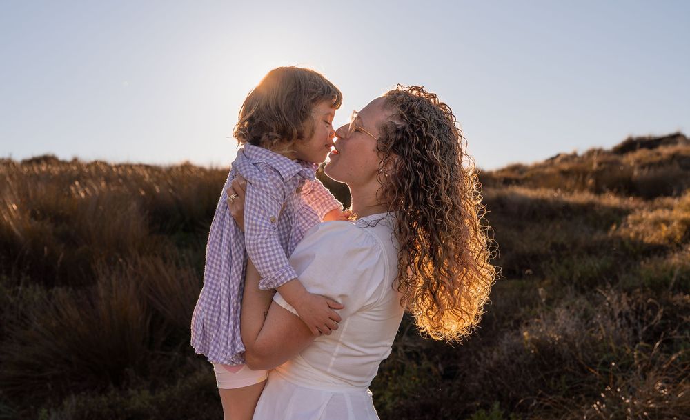 A mother in a white dress holds her toddler close, sharing a tender nose-to-nose moment in a sunlit field.