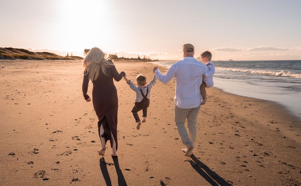 Family walking on a sunny beach at sunset, parents swinging young child between them while carrying a toddler.