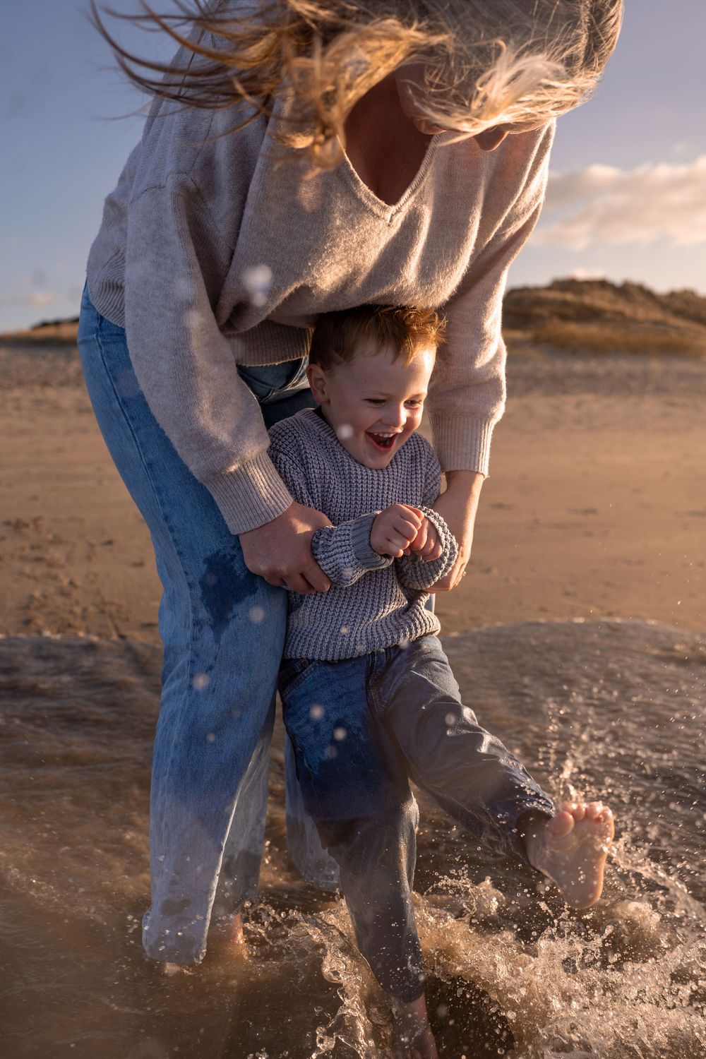 A joyful toddler laughs as a woman lifts them over splashing waves on a sunny beach.