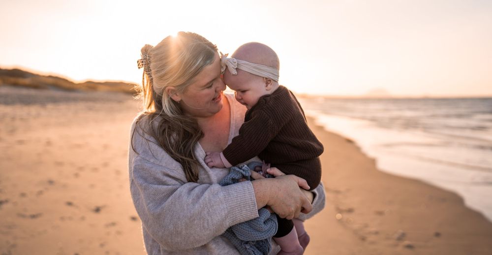 Mother holding baby on a sunny beach at golden hour, sharing a tender moment by the ocean shore.