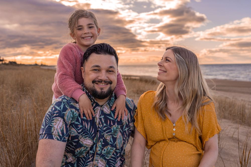 Family of three posing at sunset on a beach, parents smiling with young child sitting on dad's shoulders.