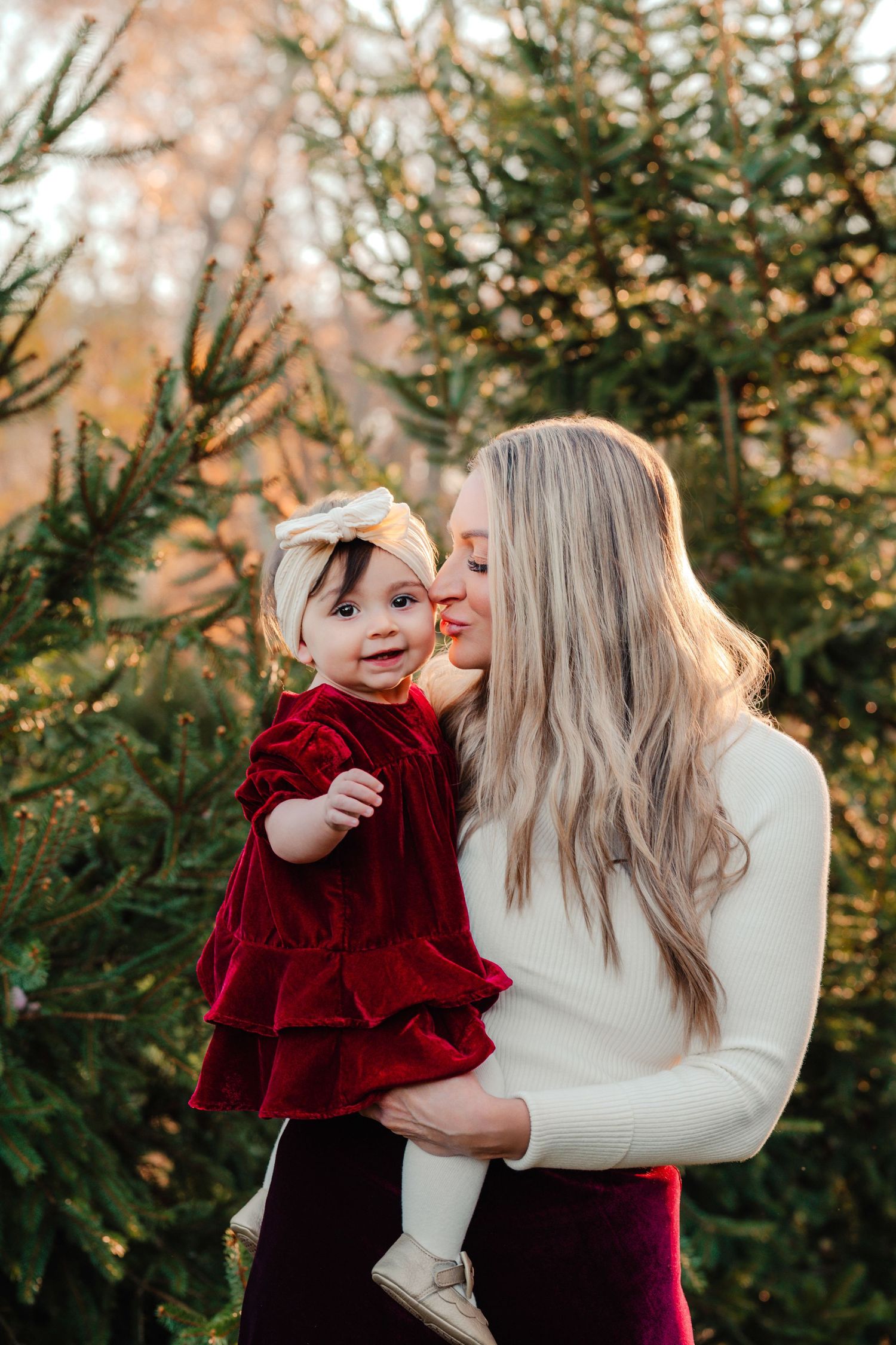 A blonde woman in a white sweater holds a baby girl in a red velvet dress near Christmas trees.