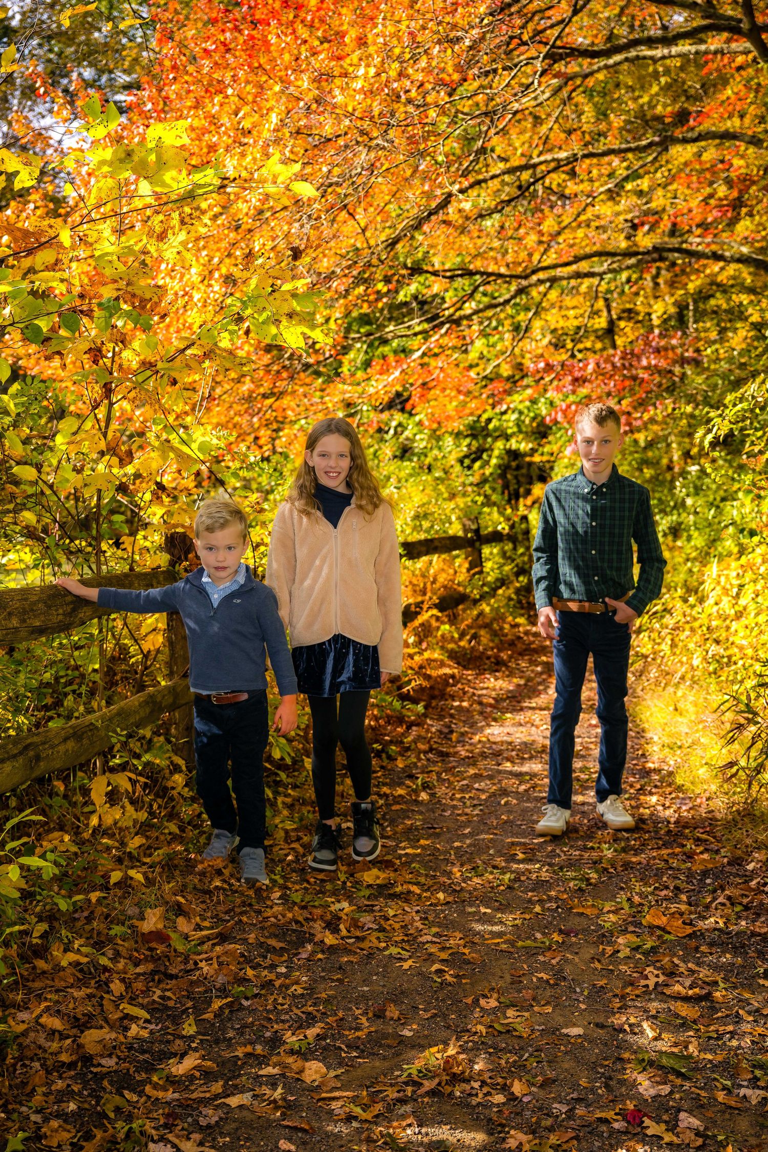 Three children pose on a leaf-covered autumn trail surrounded by vibrant red and orange fall foliage.