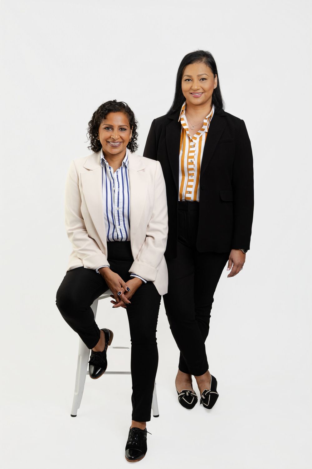 Two professional women in blazers pose confidently against a white background, one seated and one standing.