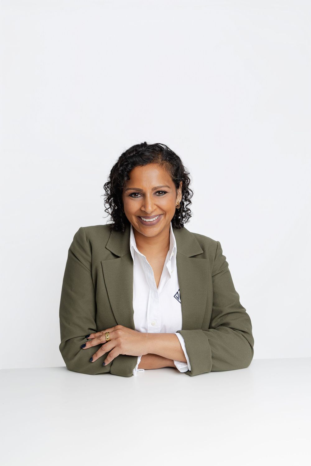 Professional woman in green blazer smiling with arms crossed at white desk, corporate headshot portrait.