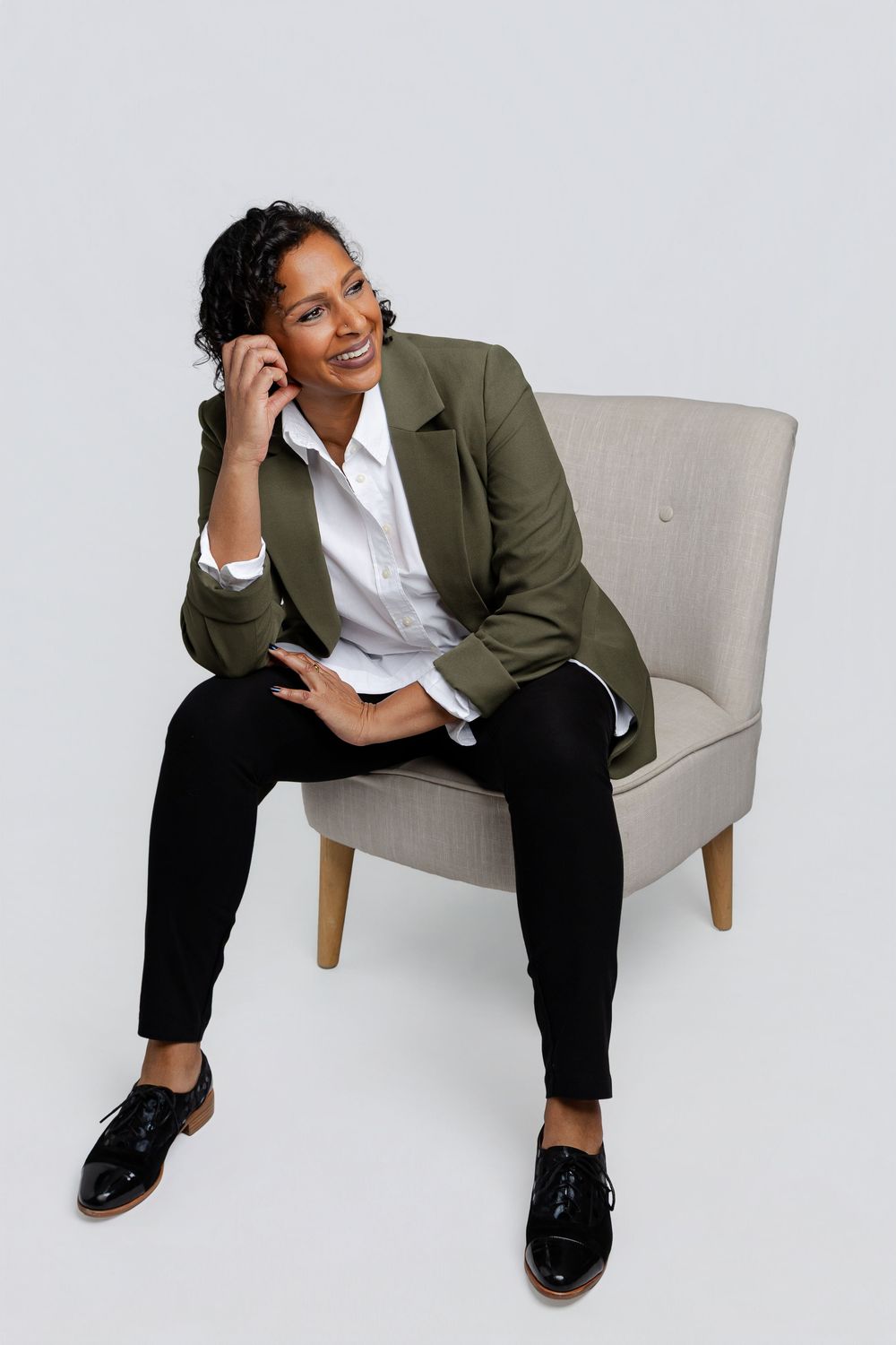 Professional woman in green blazer sitting in chair, smiling while talking on phone in studio setting.