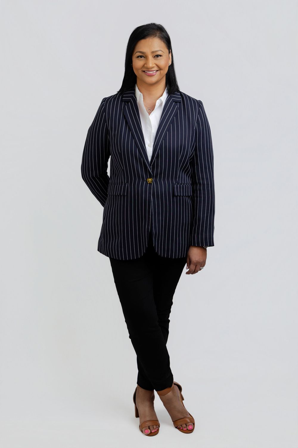Woman in navy pinstripe blazer, white shirt, black pants, and heeled sandals poses confidently against gray background.