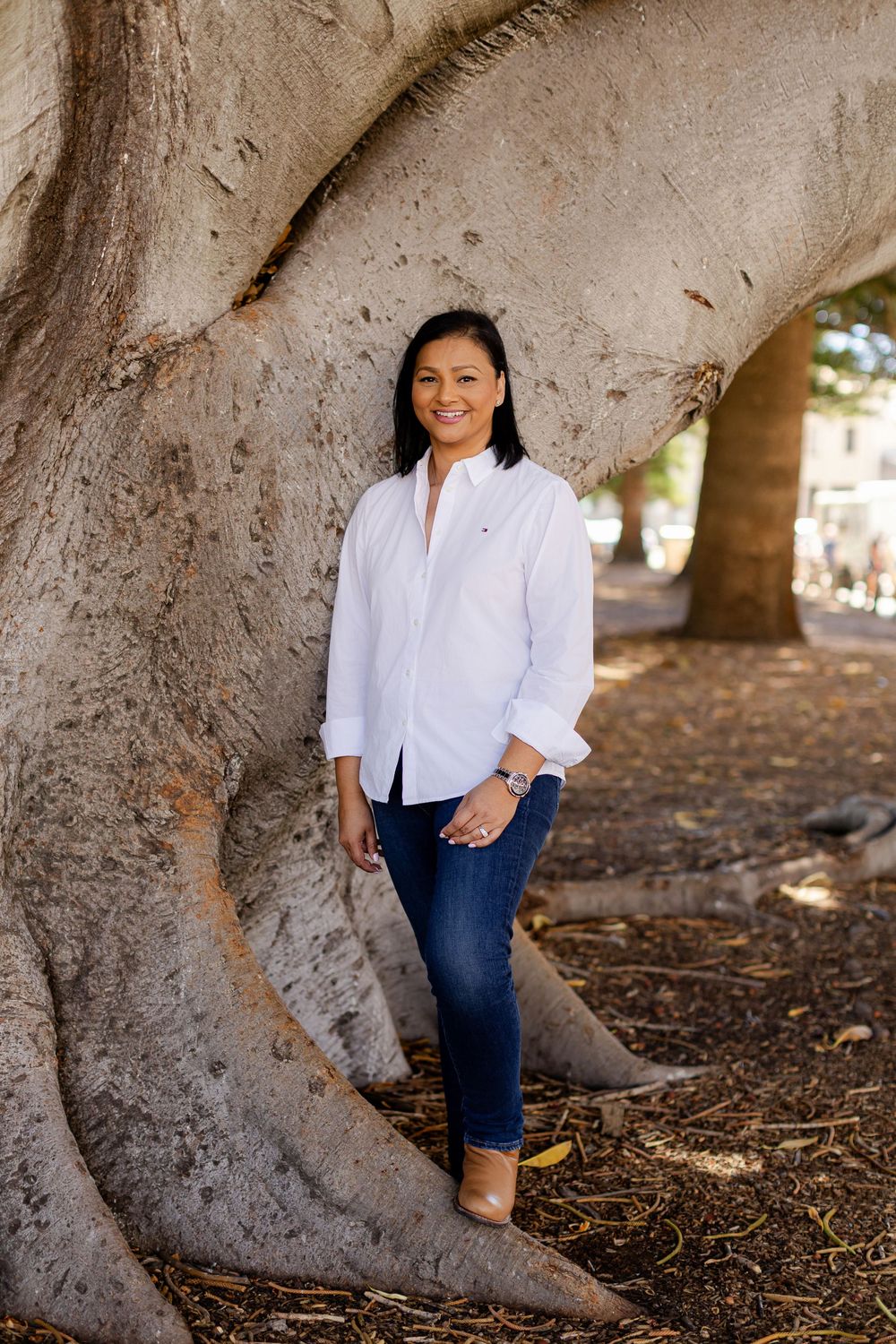 Woman in white shirt and jeans smiling beside a large tree trunk outdoors.