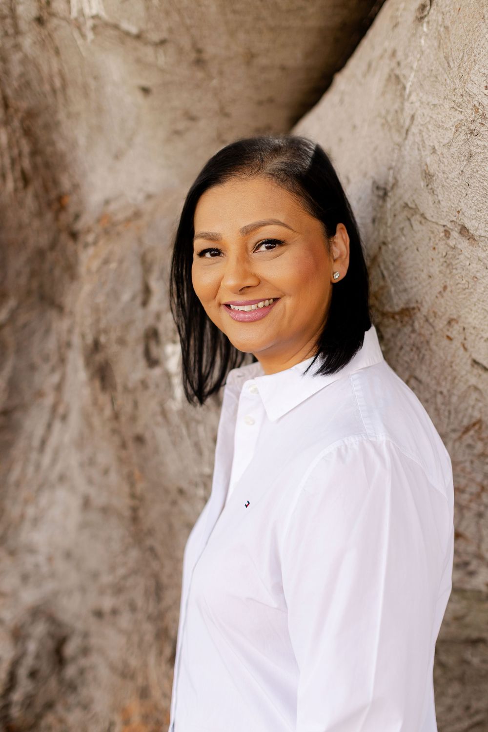Smiling woman with black hair in white button-up shirt leaning against a stone wall outdoors.